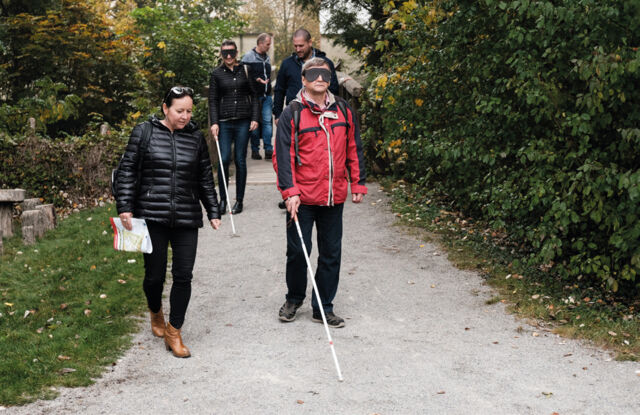 Auf dem Bild sind zwei Menschen mit Blindenstöcken und drei Begleitpersonen zu sehen. Das sind die Projektteammitglieder. Sie probieren es aus, was es heißt, mit Blindenstock unterwegs in der Natur zu sein.