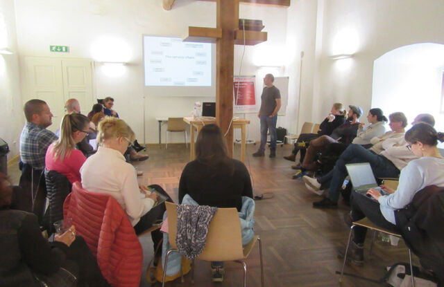 This picture shows people listening to a lecture. The room is medium-sized and divided by a column. The lecture is about service chain in a nature park.