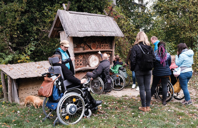 Dieses Bild zeigt Mitglieder des Projektteams beim Ausprobieren, wie sich das Naturerleben im Rollstuhl bzw. mit einem Blindenstock anfühlt. Die Begleiterin im Rollstuhl führt die Gruppe.